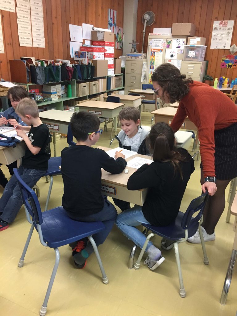 Étudiante dans une classe, penchée vers des enfants faisant des exercices sur une feuille assis à leur bureau.
