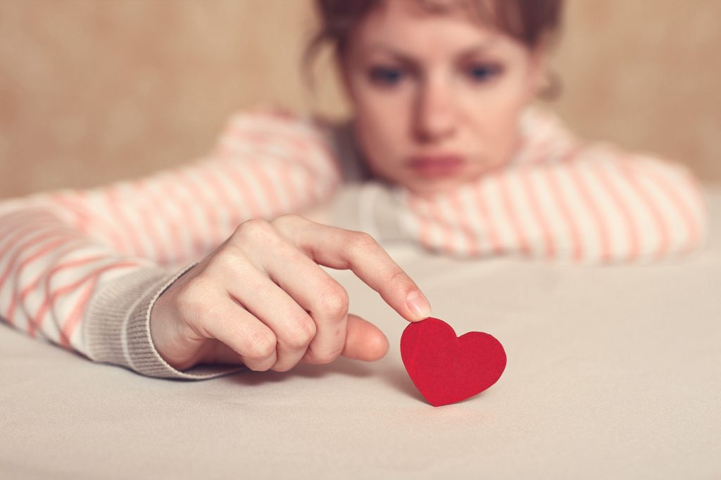 Une femme accotée sur son bras, tenant un coeur en papier, le regard triste.
