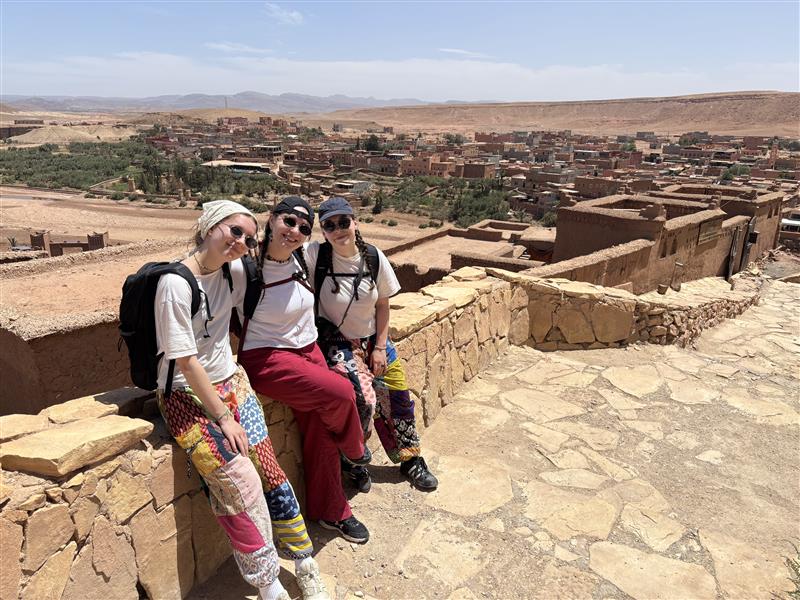 Trois étudiantes assises sur un muret de pierre, au Maroc. À l'arrière-plan, on voit au loin des édifices.
