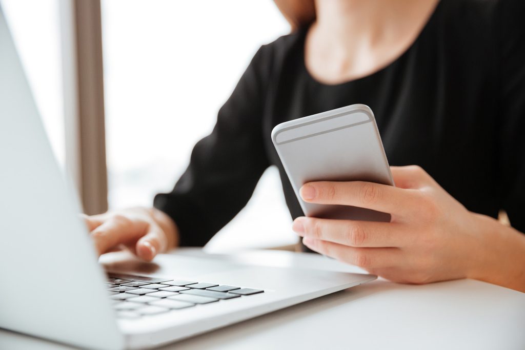 Personne en train de taper à l'ordinateur portable, avec un cellulaire dans l'autre main