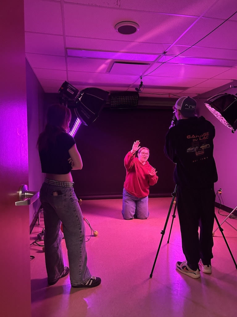 Une étudiante à genoux dans un studio, sous une lumière rosée, qui se fait filmer par deux autres personnes.