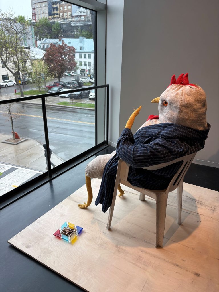 Oeuvre en textile représentant un coq portant un veston, assis sur une chaise devant une fenêtre.