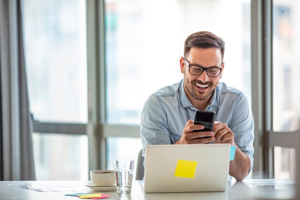 Homme avec des lunettes assis à la table de bureau devant l'ordinateur portable, tenant un téléphone portable.
