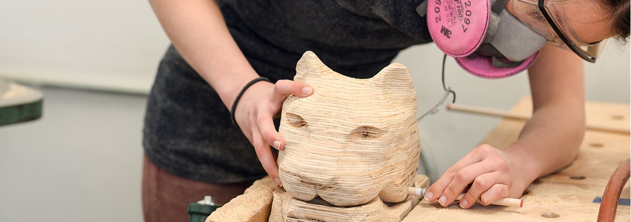 Femme avec masque rose travaillant sur une sculpture en bois ressemblant à une tête d'un animal