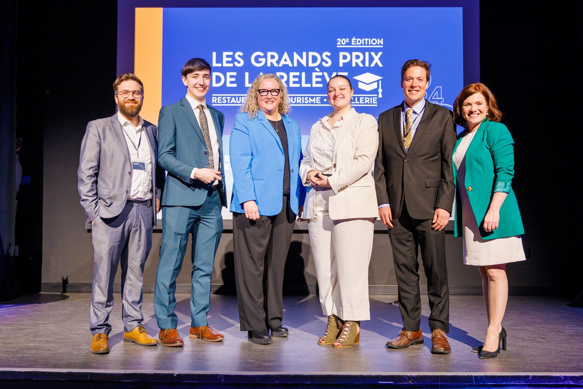 Samuel Gosselin, membre de l'Association hôtelière de la région de Québec (AHRQ), Mathieu Morissette, étudiant, Sandra Waite, enseignante en Techniques de gestion hôtelière, Raphaëlle Gindre, étudiante, Alupa Clark, directeur général de l'AHRQ, et Véronyke Tremblay, directrice générale de l'Association Hôtellerie du Québec, sur scène devant l'affiche des Grands prix de la relève.