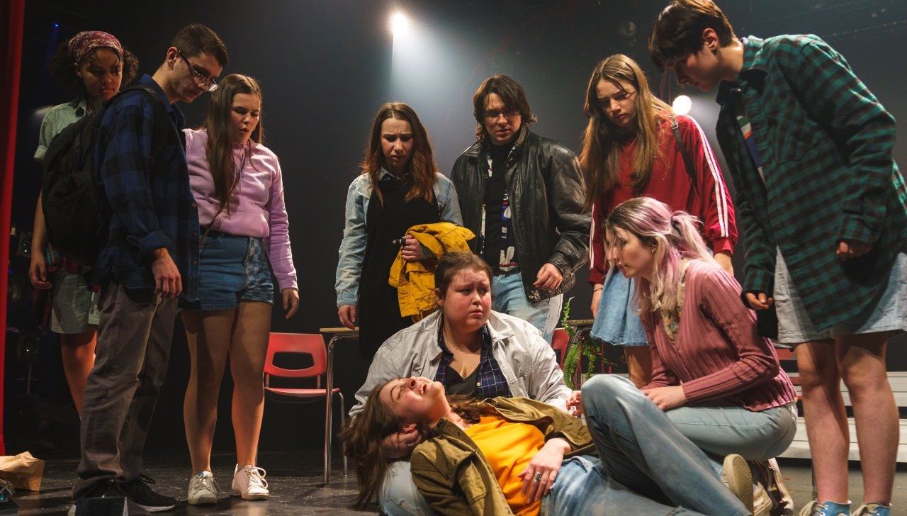 Un groupe d'étudiants de la troupe de théâtre du Grand escalier, au Cégep Limoilou, entourant l'air inquiet une personne couchée par terre, les yeux fermés et la bouche ouverte